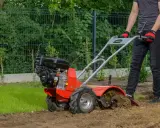 Mężczyzna w rękawiczkach obsługuje czerwoną glebogryzarkę, która spulchnia ziemię. Widać, jak działa glebogryzarka, przygotowując grunt.