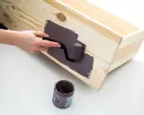 En hand använder en liten roller för att måla en träkista med lila färg, måla utan penseldrag för en jämn yta.