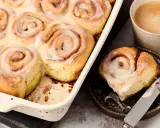 Saftige, vegane Zimtschnecken mit cremiger Glasur in einer Auflaufform, daneben eine Tasse Kaffee und eine einzelne Schnecke auf einem Teller.