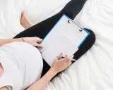 Kobieta w ciąży leży na łóżku z notatnikiem i długopisem, zastanawiając się, czy na macierzyńskim można pracować.