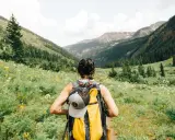 Kobieta z plecakiem wyrusza na trekking przez łąkę pełną kwiatów, w kierunku górskich szczytów.