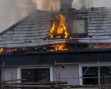 Un **incendie habitation** ravage le toit d'une maison, avec des flammes vives et une épaisse fumée s'échappant des combles.