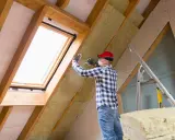 Mann isoliert ausgebauten Dachboden mit Dämmmaterial. Ein Dachfenster sorgt für Licht.