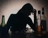 Sylwetka kobiety z kieliszkiem w dłoni, otoczona butelkami. Widoczny problem z przedawkowaniem alkoholu.