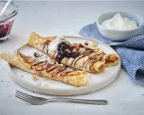 Matiga pannkakor rullade och toppade med chokladsås, blåbär och grädde. Serveras med en skål bärkompott och en blå rutig handduk.