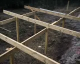 Grundstomme för att bygga altan tar form. Träbjälkar monteras på stolpar i en skogsmiljö.