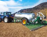 Niebieski traktor ciągnie przyczepę z cysterną do przewozu gnojowicy, wyposażoną w rozdzielacz z wieloma wężami do precyzyjnego nawożenia pól.