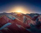 Bunte Schichten aus Rot, Gelb und Blau formen die **Regenbogenberge Chinas** unter einem tiefblauen Himmel mit aufgehender Sonne.