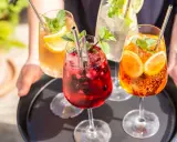 Vier erfrischende Hugo Spritz Cocktails mit Beeren, Zitrusfrüchten und Minze auf einem Tablett.