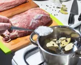 Przygotowanie solanki do wędzenia. Łyżka soli trafia do garnka z wodą, liśćmi laurowymi i pieprzem, obok surowego mięsa.