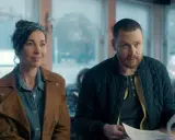 En kvinna med scarf och en man med skägg sitter vid ett bord. De ser ut att vara med i en dansk serie på SVT.