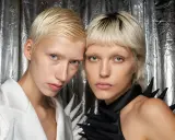 Zwei Models mit Pixie Cut feinem Haar und auffälligen Outfits vor silberner Kulisse.