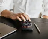 Dłoń mężczyzny nad kalkulatorem, obok laptop i długopis. Czy studia podyplomowe dają status studenta?