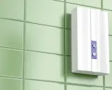 Moderner, weißer Durchlauferhitzer ohne Neutralleiter an einer hellgrünen Fliesenwand. Zwei Drehregler zur Temperatureinstellung.