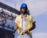 Chief Keef, w niebieskiej czapce i żółtej koszuli z kwiatami, na scenie. Jego wiek nie jest widoczny, ale styl mówi wszystko.