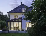 Moderner Anbau Wintergarten mit Glasfront und Beleuchtung erweitert ein Haus. Der Garten ist abends beleuchtet.
