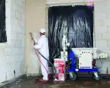 Pracownik w białym kombinezonie natryskuje tynk na ścianę z bloczków. Obok stoi maszyna m-tec M300 i worek z napisem "lekkie tynk".