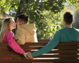 Kobieta w różowej bluzce całuje się z mężczyzną w beżowej bluzie, podczas gdy trzeci mężczyzna siedzi obok, trzymając ją za rękę. Zdrada w parku.