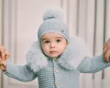 Un tierno bebé con un conjunto de punto azul claro, gorro y abrigo con cuello de pelo. Sus manos son sujetadas por adultos, como si estuviera aprendiendo a caminar. Este patrón de pelele para bebé es adorable.