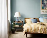 Dormitorio con paredes azul claro y ropa de cama a rayas amarillas y blancas. Los relajantes colores para dormitorios se complementan con una mesita de noche de mimbre y un jarrón con flores.