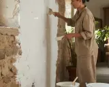Une femme en tenue de lin est en train de peindre à la chaux un mur rustique. Des plantes vertes décorent l'arrière-plan.