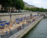 Menschen entspannen sich am **Paris strand** unter blauen Sonnenschirmen. Palmen und Gebäude säumen die Uferpromenade.