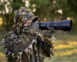Fotograf w ghillie suit z aparatem do fotografowania zwierząt czeka na idealny kadr.