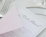 Eleganckie zaproszenie z osobą towarzyszącą, "You're Invited", na różowej kopercie, obok sztućców i talerza.