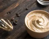 Cremige Crema al Caffè in einer Tasse, daneben ein Handmixer mit Teigresten und Kaffeebohnen auf Holz.