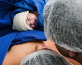 Noworodek w czepku, otulony niebieskim materiałem, leży na brzuchu mamy. Widoczna twarz dziecka i fragment twarzy mamy. To piękny moment porodu lotosowego.
