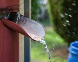 Agua de lluvia cayendo de una canaleta roja hacia un barril azul, una forma práctica de saber como recoger agua de lluvia en un terreno.