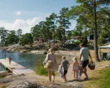 Familj på väg mot vattnet för roliga aktiviteter barn. Soligt, skärgårdsmiljö med röda stugor och camping.