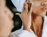 Une femme applique une crème pour l'hydratation du visage, enveloppée dans une serviette et un peignoir douillet.