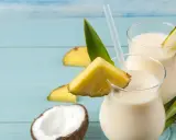 Dwa orzeźwiające koktajle Pina Colada, ozdobione kawałkami ananasa i liśćmi, na tle niebieskich desek. Idealny przepis na lato!