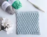 Muestra de tejido a crochet con el patrón nudo de salomon en color azul claro, junto a ovillos de lana gris y verde, una flor blanca y un ganchillo.