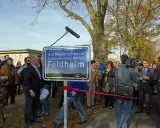 Feldheim, ein Vorreiter unter den energieautarken Gemeinden Deutschlands, wird hier mit einem Schild gefeiert. Eine Gruppe von Menschen, darunter ein Kameramann, versammelt sich.