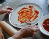 Ręce rozprowadzają pyszny sos do pizzy na cieście. Obok leży wałek i miseczka z sosem.