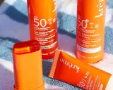 Trois produits solaires orange vif, dont un stick, sur une serviette rayée. Idéal pour savoir quelle crème solaire choisir pour une haute protection SPF 50.