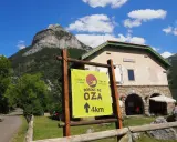 Cartel indica "Bosque de Oza 4km" en un día soleado en el valle de hecho, con una casa forestal y montañas al fondo.