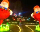 Gigantesche lanterne di bambini paffuti illuminano un sentiero a Wonderland Rimini, con due persone che salutano tra fiori luminosi.