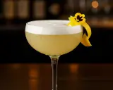 En elegant elderflower sour med skummig topp, garnerad med en citronskal och en liten blomma.