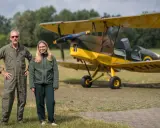 Mann in Fliegerkluft und junge Frau stehen neben gelbem Doppeldeckerflugzeug. Das Flugzeug erinnert an einen Tiger Moth.