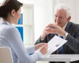 Kobieta i starszy mężczyzna rozmawiają przy biurku. Ona pokazuje mu CV, szukając pracy dodatkowej z domu.