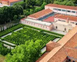 Un giardino con un intricato labirinto borges, circondato da edifici con tetti rossi e alberi verdi.