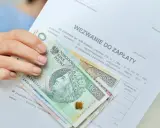 Dłoń trzyma wezwanie do zapłaty i polskie banknoty 100 i 200 zł. Dokument zawiera informację o zawiadomieniu o uchyleniu zajęcia.