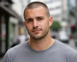 Hombre joven con corte en u, barba cuidada y camiseta gris, mirando a la cámara en una calle urbana.
