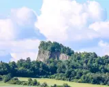 Skalisty szczyt porośnięty drzewami, przypominający wilczą górę, wznosi się nad zielonymi polami i lasami.