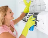 Mujer con guantes verdes limpia el aire acondicionado que huele a humedad, rociando un producto.
