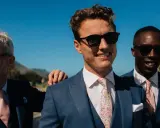 Four men in blue suits with pink ties and sunglasses, embodying a "dress to impress" dress code for a sunny outdoor event.