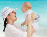 Kobieta w białym kapeluszu unosi dziecko na plaży. Radość macierzyństwa, nawet w przypadku surogacji, jest widoczna w ich uśmiechach.